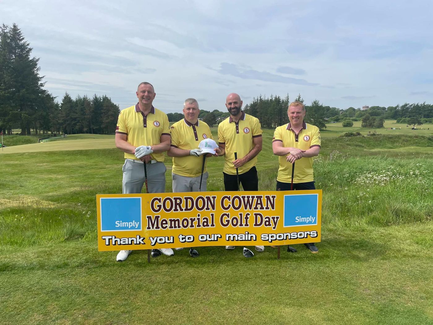 Four golfers in yellow shirts beside the Gordon Cowan Memorial Golf Day banner